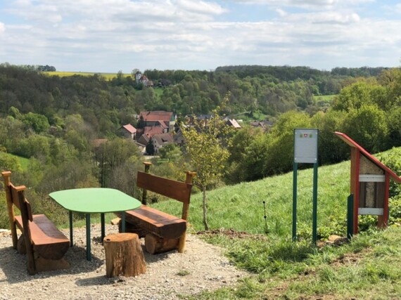 Sitzgelegenheit mit Tisch, Hinweisschild und Insektenhotel im Vordergrund. Blick über das Tal. Unten Standorf gegenüber die Ulrichskapelle.